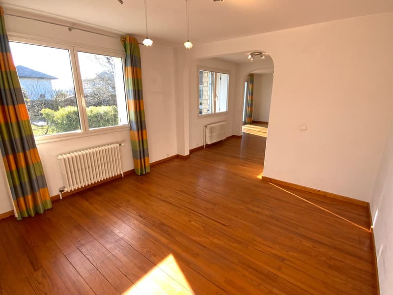 An empty room with hardwood flooring, large windows with curtains, wall-mounted radiators, and ceiling fixtures.