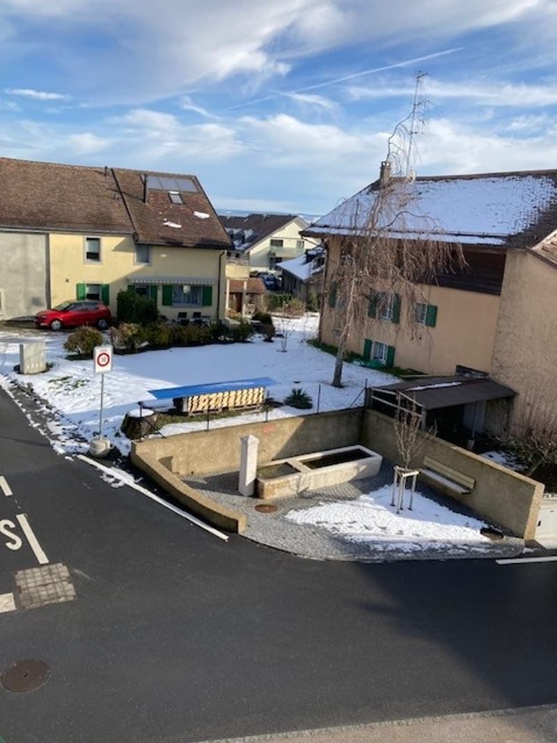 Houses with snow on the roof, red car parked in front, snow on the ground, bench, and a small fountain