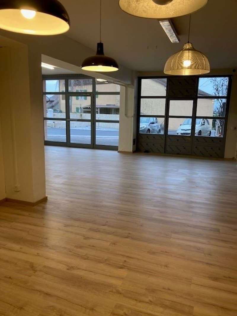 Large open space with hardwood flooring, multiple pendant lights, and large windows providing natural light.