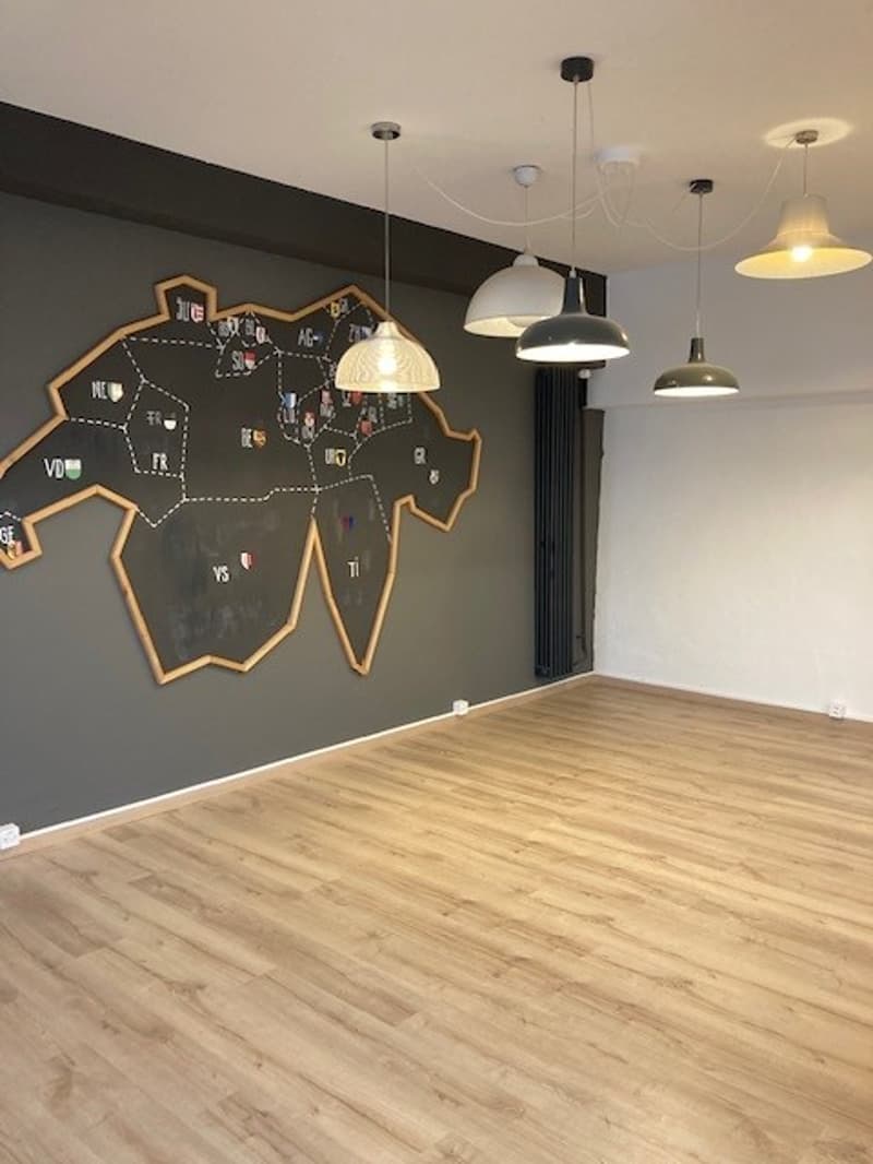 Large open space with hardwood flooring, dark gray walls, and multiple pendant lights. A large chalkboard map is displayed on the wall.