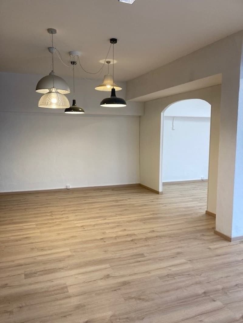 This image shows an empty room with hardwood floors, white walls, and several pendant lights hanging from the ceiling. The room appears to be a living or dining area, with an arched doorway visible in the background.