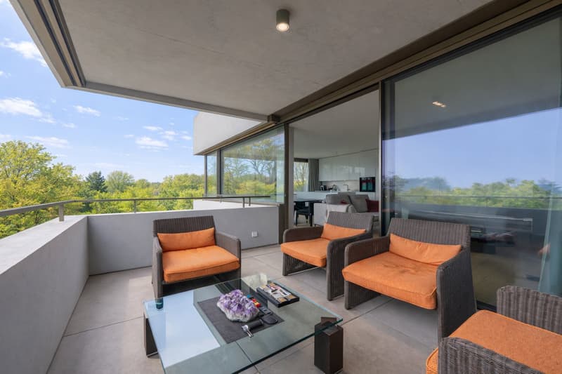 Modern balcony with large glass doors, gray walls, white ceiling, tiled floor, couch, glass coffee table, orange cushioned chairs, trees view