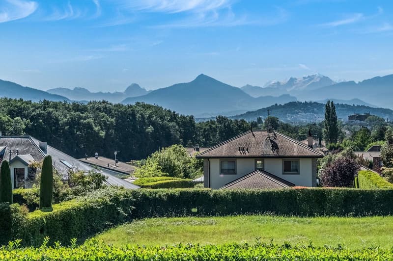 The image shows a residential area with houses surrounded by lush greenery and trees. In the background, there are mountains and a scenic landscape with a blue sky. The houses have tiled roofs and appear to be in a suburban or rural setting.
