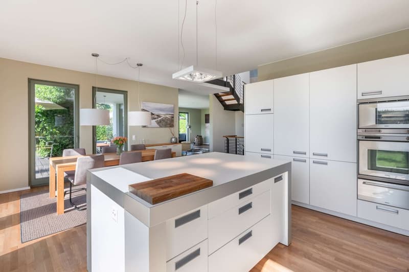 Spacious open-concept kitchen with white cabinets, wooden countertops, and stainless steel appliances including a dishwasher. The kitchen flows into a dining area with a wooden table and chairs. Large windows provide natural light and views of the surroun