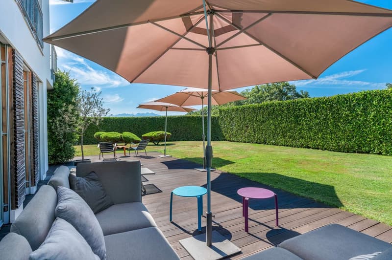 Spacious outdoor patio with a large beige umbrella, comfortable gray sofa, colorful stools, and a well-manicured lawn surrounded by a hedge. The area appears to be suitable for relaxation and outdoor entertainment.