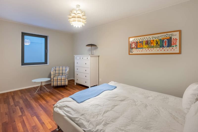 Spacious bedroom with hardwood flooring, large window, white walls, modern light fixture, dresser, and armchair