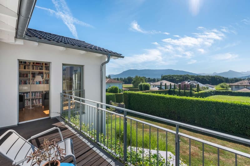2-story house with a balcony overlooking a scenic landscape, including a well-manicured garden, hedges, and distant mountains. The house has a bookshelf visible through the window, indicating a study or library area.