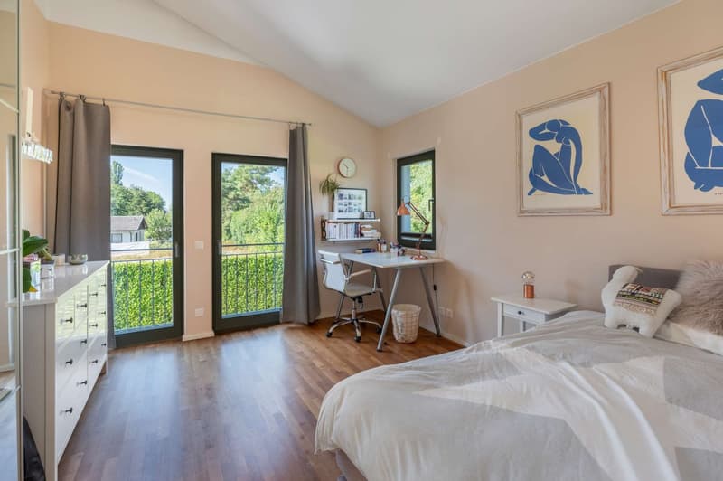 Spacious bedroom with hardwood floors, large windows providing natural light, a desk area, and artwork on the walls. The room has access to a balcony or terrace overlooking the surrounding greenery.