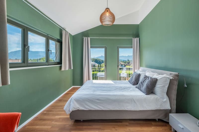 Spacious bedroom with green walls, large windows offering a scenic mountain view, wooden flooring, a king-size bed with gray bedding, and a decorative pendant light fixture.