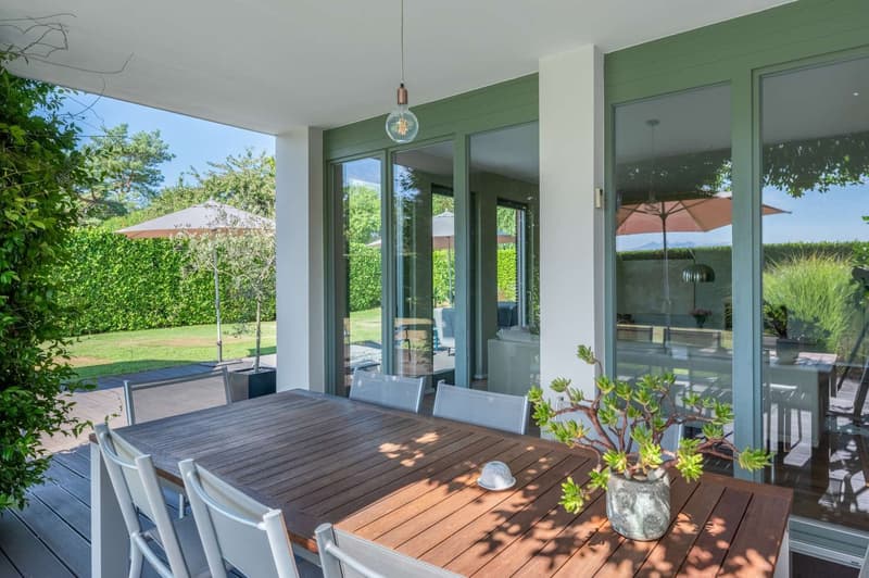 Covered outdoor dining area with a wooden table and chairs, surrounded by large windows overlooking a lush green garden. The space features a pendant light fixture and potted plants, creating a cozy and inviting atmosphere.