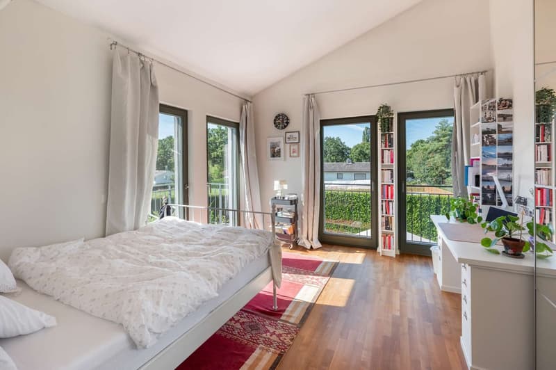 Spacious bedroom with large windows, hardwood floors, white bedding, and a balcony overlooking the greenery outside. The room also features a bookshelf, potted plants, and decorative wall art.