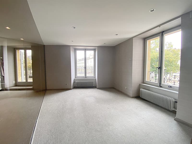Empty room, carpeted floor, two windows, two radiators, doors to other rooms.