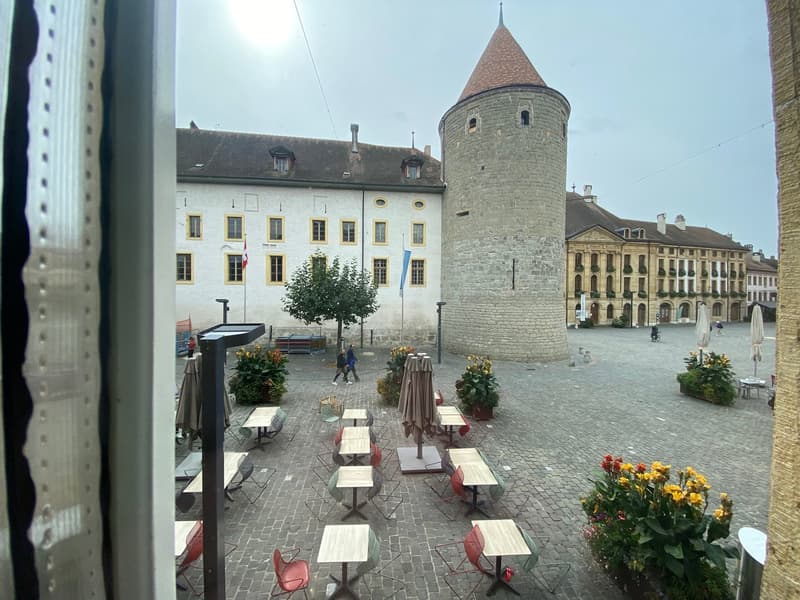View of a historical building with a tower, outdoor seating area with tables and chairs, flag poles, flowers, and cobblestone pavement.