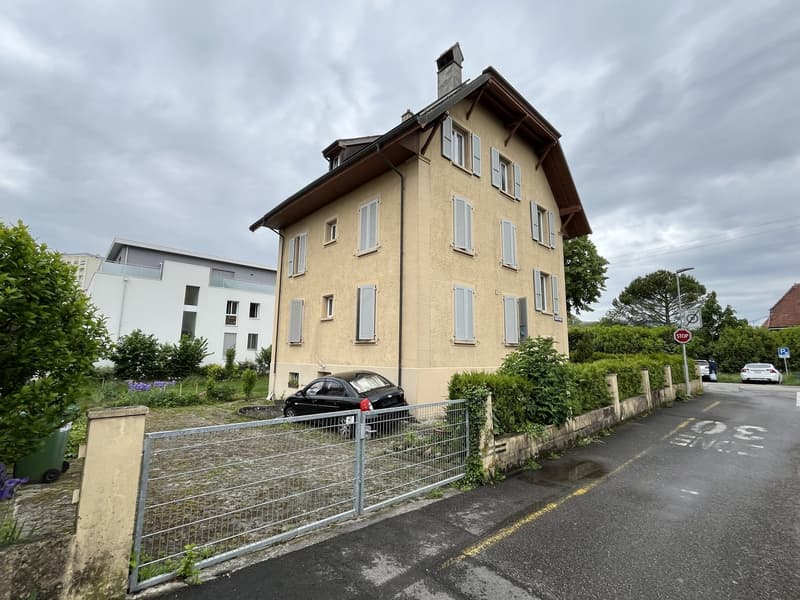 Multi-story building with beige walls, multiple windows, chimney, car parked in front of garage
