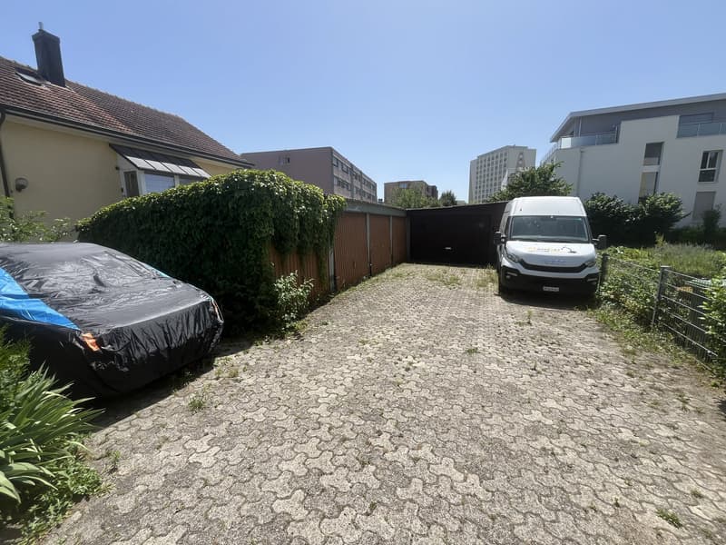 Empty driveway with a van, house on the left with brick roof and chimney, another house on the right with multiple floors, fenced with a gate