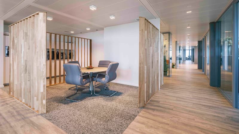 The image shows a modern office space with a conference room. The room has a wooden table and chairs, and the walls are made of wooden slats. The hallway outside the conference room has a wooden floor and glass walls. The space appears to be well-lit with