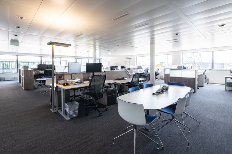 Large open office space with multiple workstations, computers, and a conference table. The space has high ceilings, large windows, and modern office furniture and equipment.