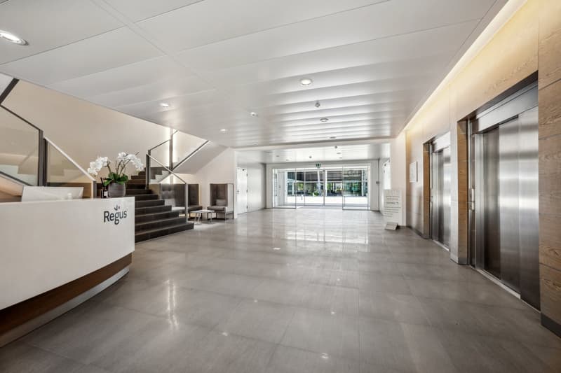 The image shows the interior of a modern office building. The lobby area features a large, open space with a high ceiling, polished tile flooring, and a reception desk with the Regus logo. The space has a clean, minimalist design with neutral colors and p