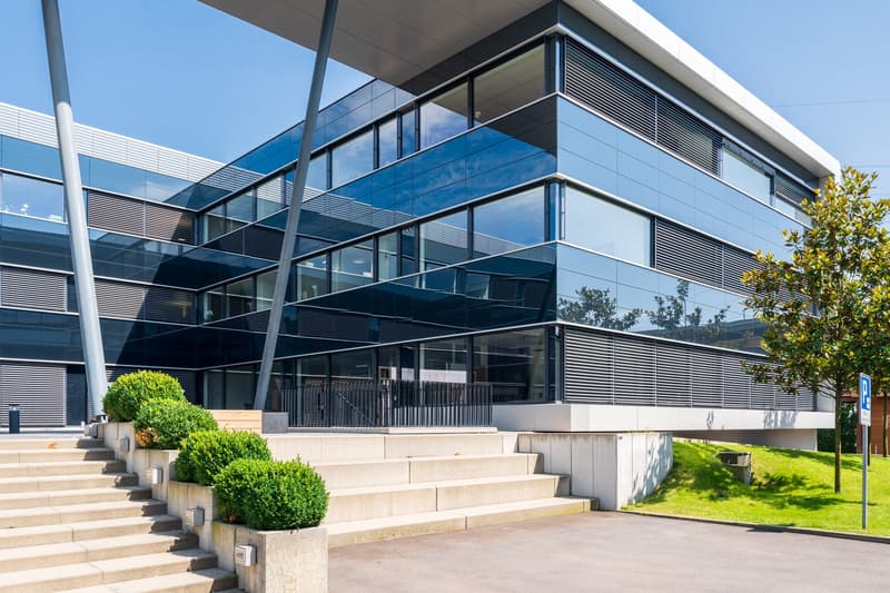 Modern multi-story office building with glass facade, balconies, and landscaping around the entrance steps.