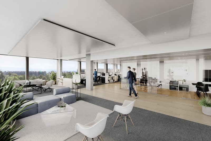 Spacious open-plan office with large windows offering a view of the city skyline. The space features a lounge area with comfortable gray sofas, white chairs, and potted plants. There is also a separate work area with desks and computers, as well as a glas