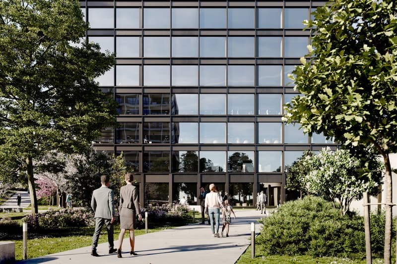 A modern office building with a glass facade surrounded by trees and greenery. People are walking on the path in front of the building.