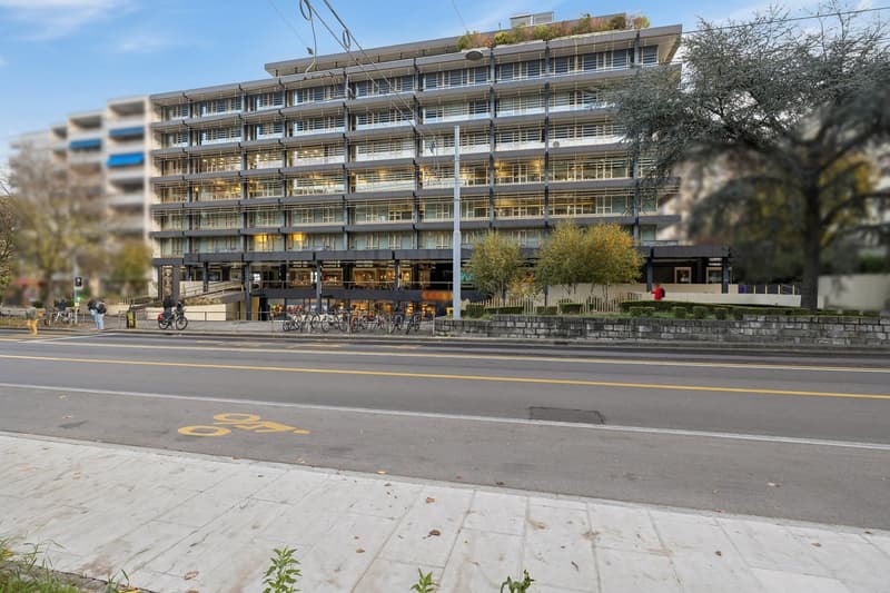 a large multi-story building with many windows, bicycles parked in front, street lamps, a yellow bike symbol on the road