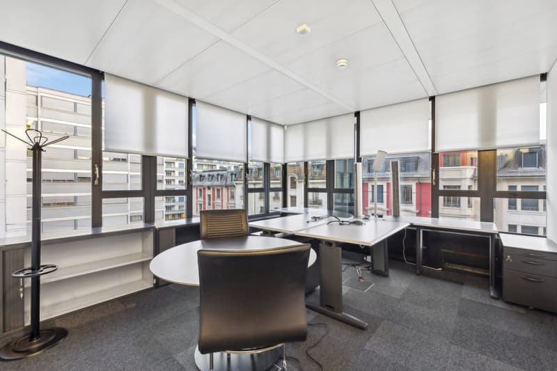 Office with large windows, multiple desks, modern furniture, ceiling lamps, gray flooring, exterior view of buildings
