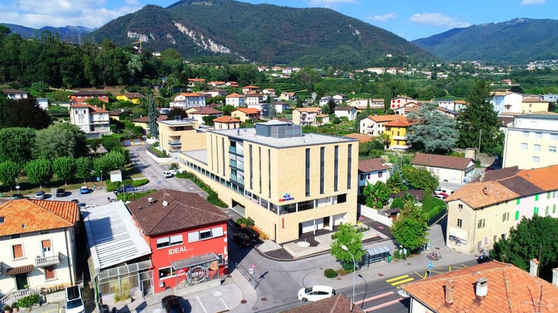 Mountain in the background, several houses of different styles, a building with a sign 'Paleomedica', multiple cars parked in front of houses, street lights, and bushes. Two cars are parked on the side of the road, and a bench is placed near the trees. A