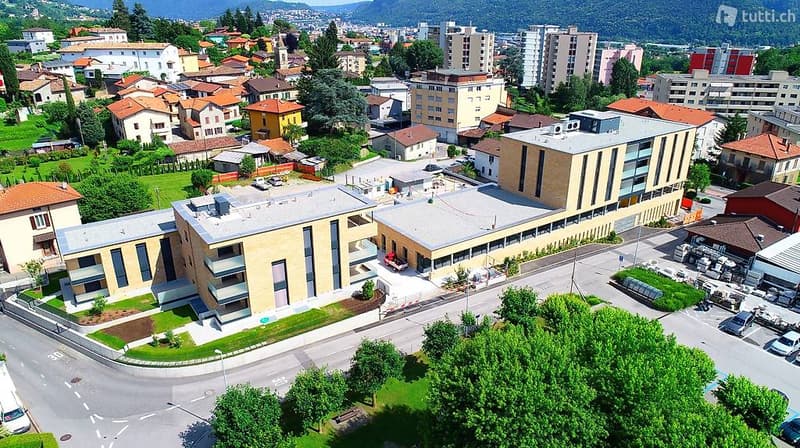 Multi-story building, beige and brown facade, large windows, nearby residential buildings, green landscape, cars parked in the lot