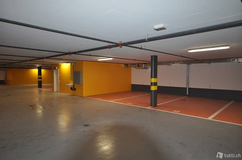 Large underground parking area with bright lighting, white and orange painted walls, grey floor, and poles, clean and empty.