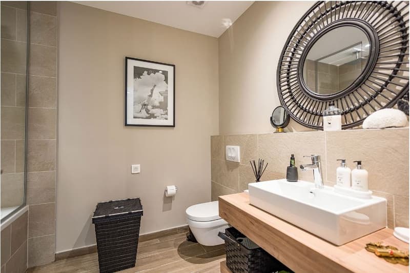 contemporary design bathroom with beige tile, a large mirror, a white sink with a faucet, a white toilet, a picture frame above the toilet, and a mirror with a wooden frame above the sink