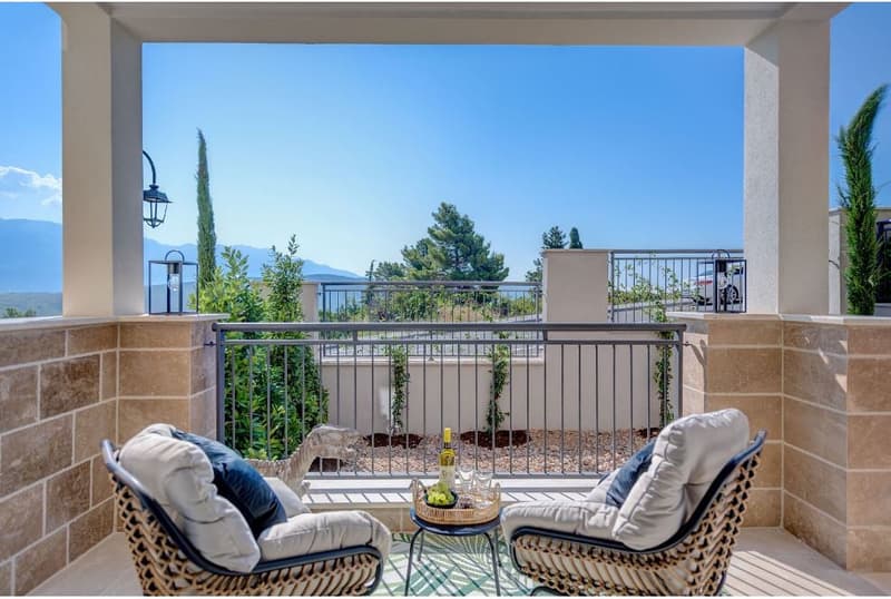 two wicker chairs with cushions, small table with wine bottle and grapes, railings, sunny view of mountains