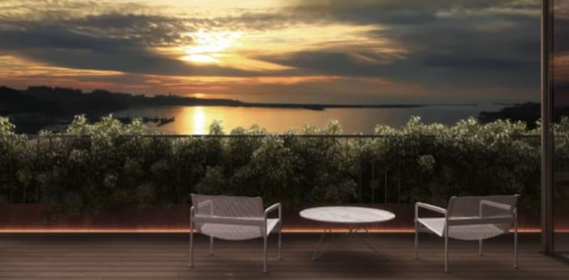 The image shows an outdoor patio or terrace overlooking a scenic lake or body of water. The patio has a round table and two chairs, providing a comfortable seating area to enjoy the beautiful sunset view over the water. The landscape is filled with lush g