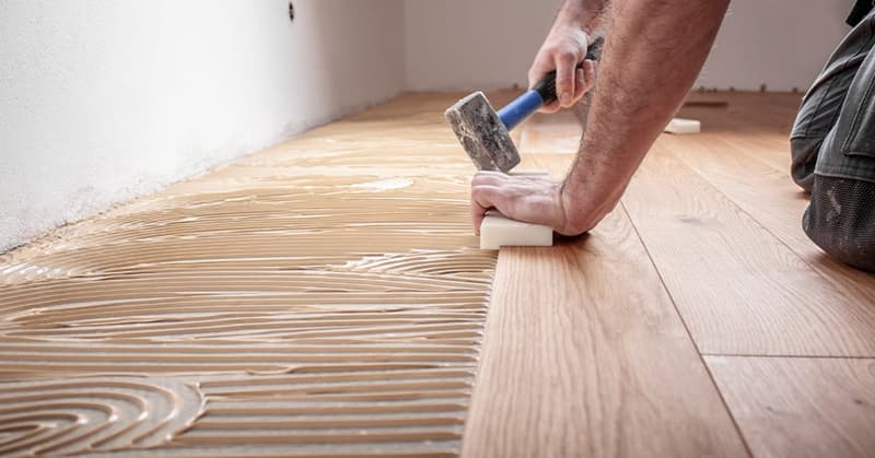 Hardwood floor being installed on concrete base, person using hammer, wall with white paint, grooves for installation