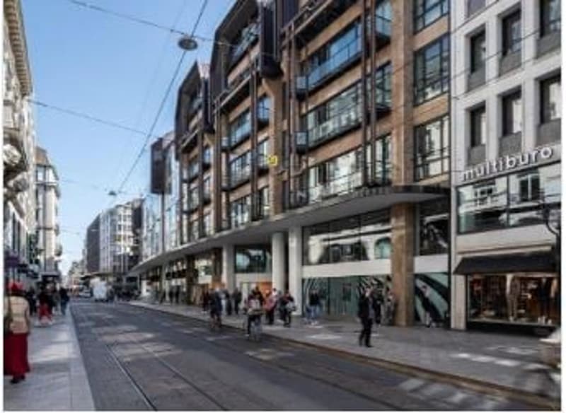 Busy city street with tram tracks, multi-story buildings with shops and restaurants, pedestrians walking on the sidewalk.