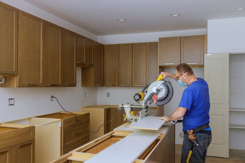 Unfinished kitchen, wooden cabinets, countertop, electrical outlet on wall, sawing machine, open door, man using mask, hammer on belt