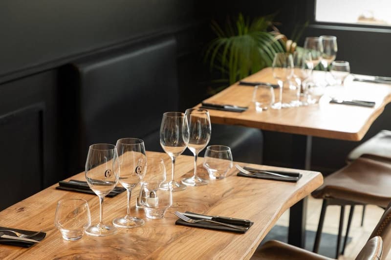 Wooden dining table set with wine glasses, cutlery, and a plant decoration. Dark walls and furniture create a cozy atmosphere. The table is set for a dining experience, with the visible features including a dishwasher, suggesting this is a restaurant sett