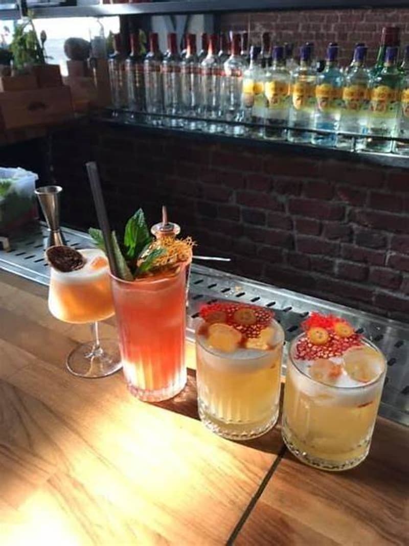The image shows a bar setting with various cocktail glasses and bottles on display. There are three cocktail drinks on the wooden bar top, each garnished with different fruits and herbs.