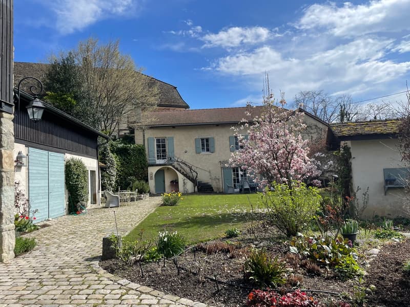Two-story stone and wood house with a garden, cobblestone driveway, and a blooming cherry tree. The house has a balcony and a garage on the side.