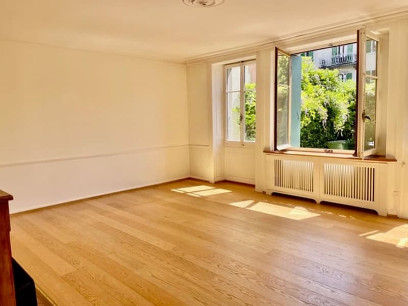 Spacious living room with hardwood floors, large windows overlooking a garden, and a radiator under the window.