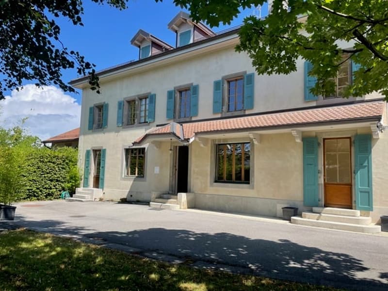 Two-story house with a beige exterior, green shutters, and a tiled roof. The house has several windows and a balcony or terrace on the upper level. The property is surrounded by trees and has a paved driveway or walkway leading to the entrance.