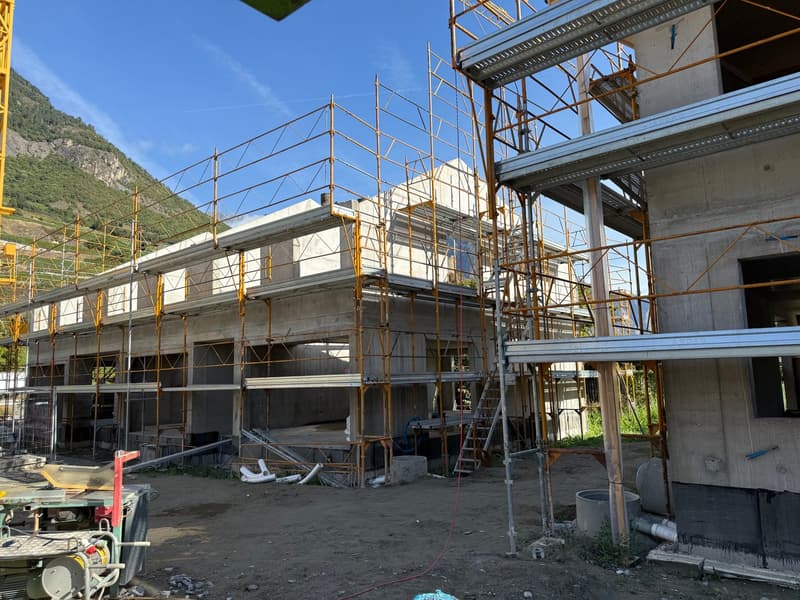 Building under construction with concrete walls, surrounded by metal scaffolding and steel beams, temporary fencing and machinery present, mountain and blue sky in the background.