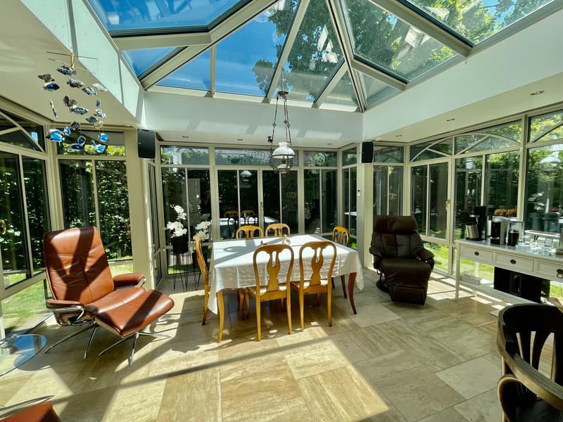 sunlit conservatory with a large glass ceiling, table and chairs, comfortable armchairs, desk with a coffee maker, and a decorative fish mobile hanging from the ceiling.