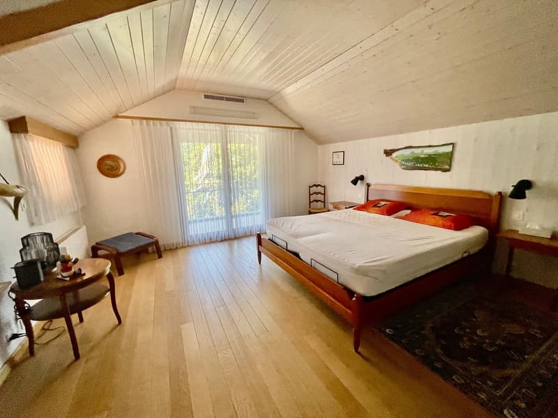 Wooden bed with two mattresses, bedside tables with lamps, rug, table with chair, wooden stool, armchair, window with curtains, artwork on the wall, radiator