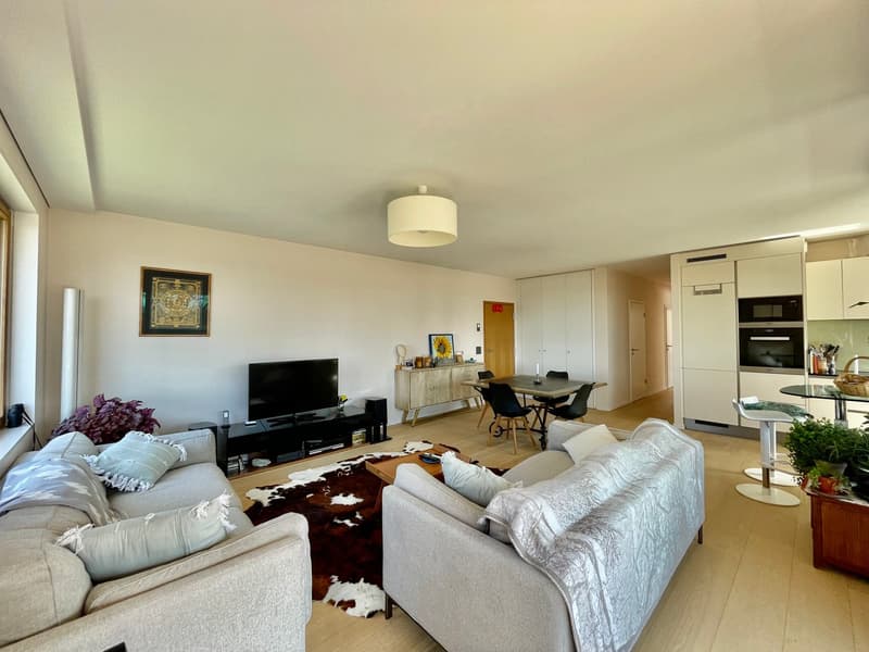Open-concept living room and dining area, with a large couch, TV stand, and dining table. The kitchen is visible in the background, with modern appliances. The room has large windows, hardwood floors, and a neutral color scheme.