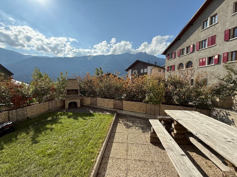 Garden: wooden table, benches, gravel path, green lawn, bushes, fireplace, wooden deck, mountains in the background.