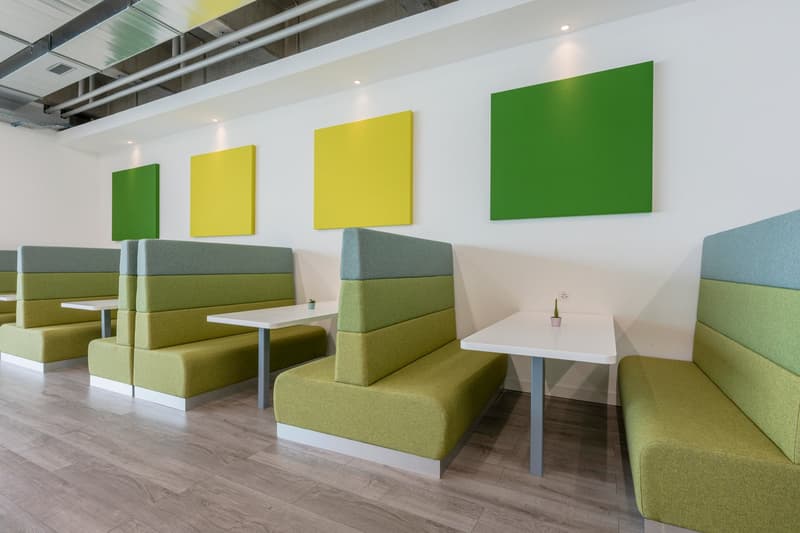 The image shows a modern and colorful office or workspace interior. The space features green and yellow accent walls, as well as comfortable green and gray seating areas with small white tables. The flooring is a wood-like laminate material. The overall d