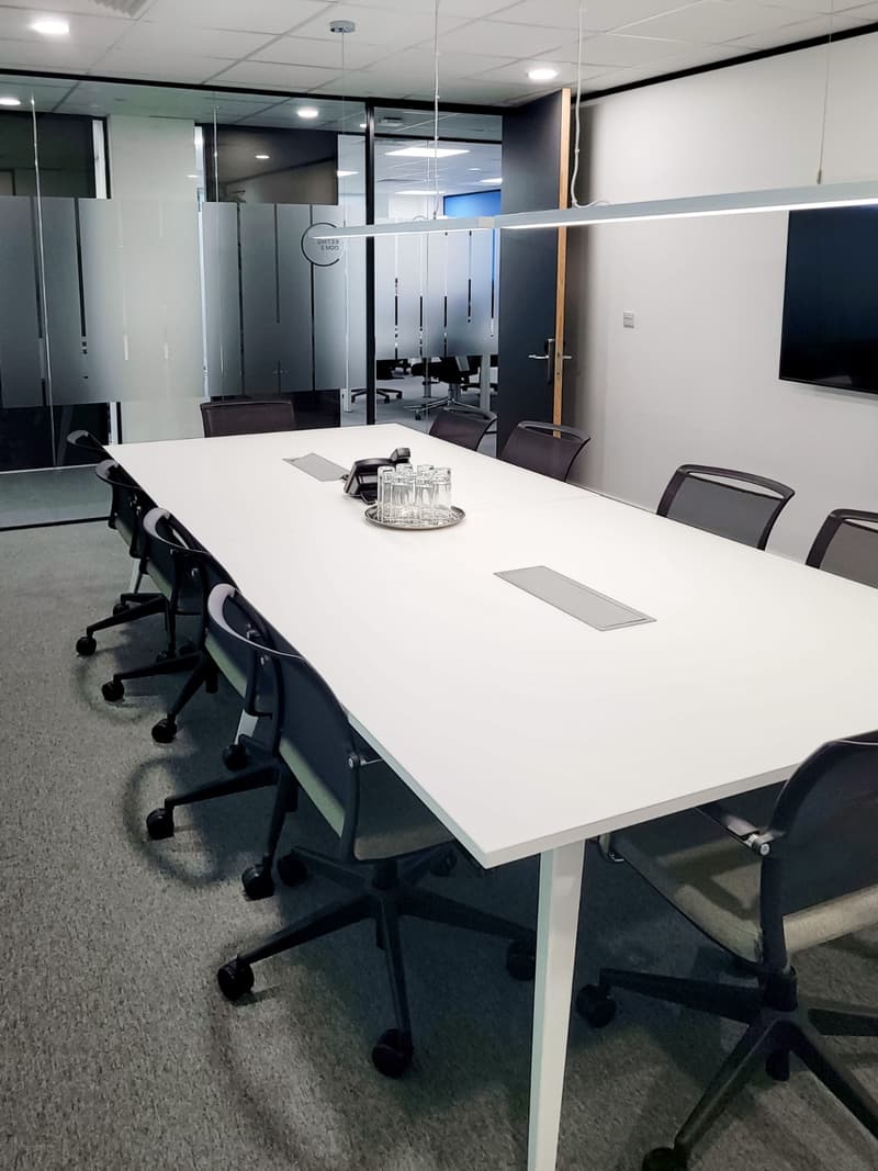 Large white conference table with black office chairs, glass walls, TV screen, and a tray of glasses on the table