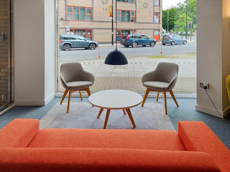 The image shows a modern and stylish interior space with large windows overlooking a street. The space features an orange sofa, two beige armchairs, a round white coffee table, and a black pendant light. The floor is covered with a gray rug, and the walls