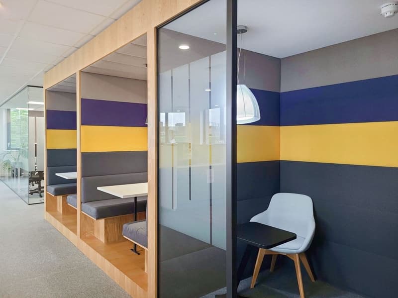 The image shows an office space with a modern and colorful design. The walls are painted in a combination of blue, yellow, and gray stripes, creating a vibrant and dynamic atmosphere. The space features glass partitions that allow natural light to flow th